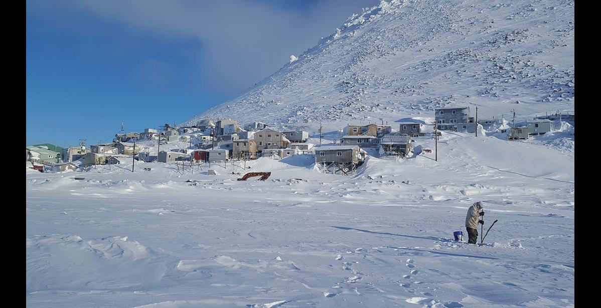 Photo by Robert Soolook LITTLE DIOMEDE— A Little Diomede man fishes through the ice in this February 2019 photo. The community of Little Diomede is shown in the background, built on the steep slopes of the island located in the middle of the Bering Strait.