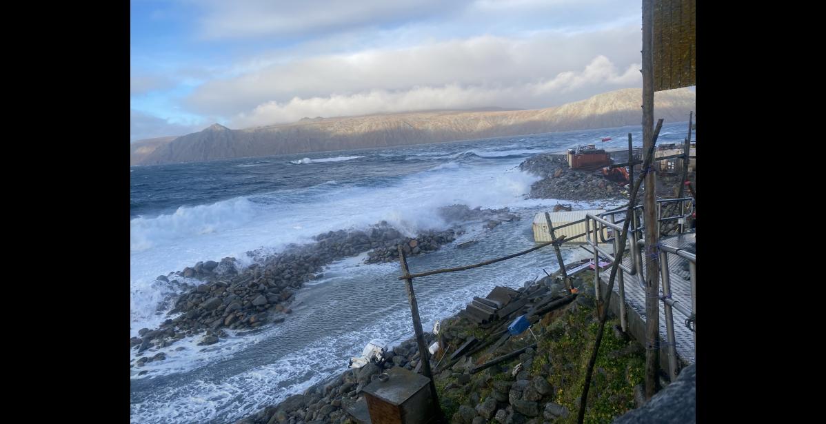 Waves crash onto the beach at Little Diomede. A container van with housing material and the tribe's excavator are both in a pond of sea water. Photo by Frances Ozenna